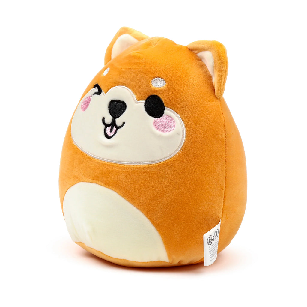 Angled view of an orange and cream Shiba Inu dog plush toy, depicting its winking face with embroidered black eyes, nose, and mouth with a pink tongue, alongside pink blush marks. A white product label is visible on its right side.