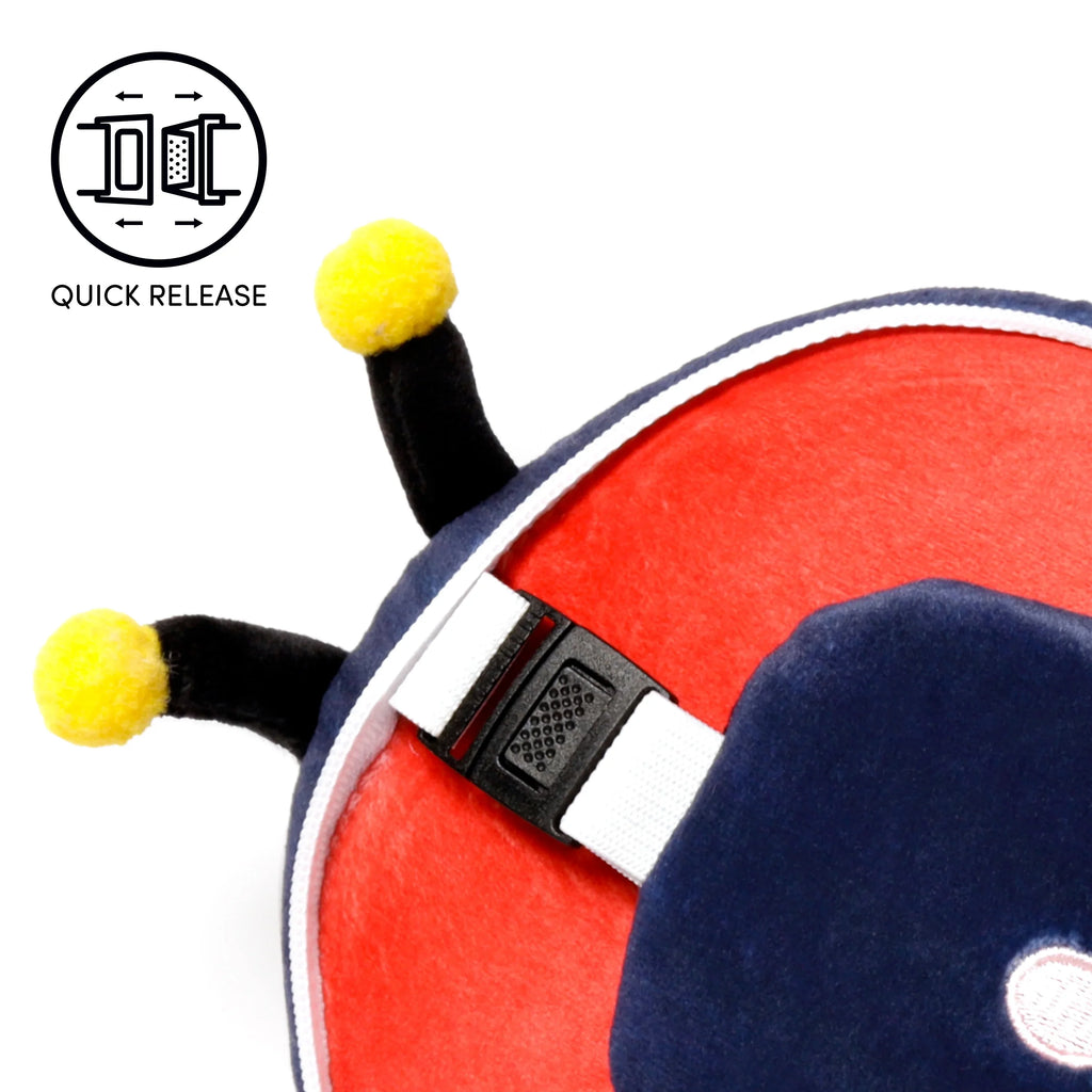 Close-up of a red and navy blue ladybug travel pillow, showing a black quick-release buckle on a white strap and two black antennae with yellow pom-pom tips. An illustrative 'QUICK RELEASE' icon appears in the top left.