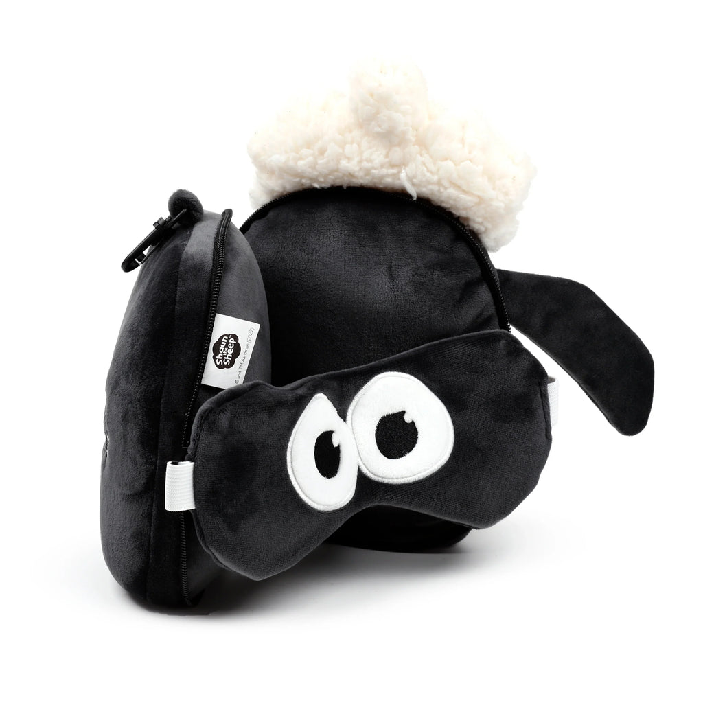 Angled view of a black plush travel pillow and eye mask set, designed as Shaun the Sheep, featuring the pillow's white fluffy wool and the eye mask's embroidered white and black eyes.