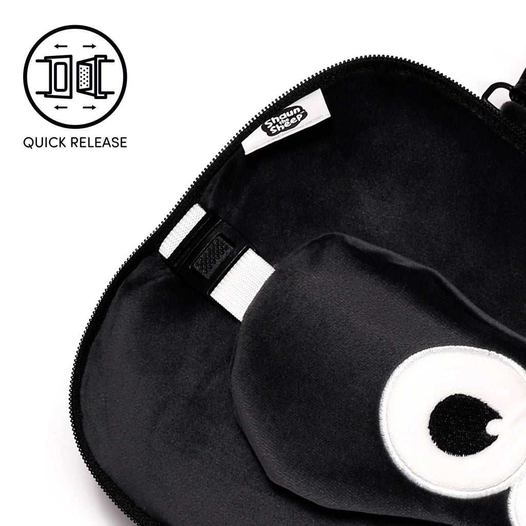 Angled close-up view of a black plush travel pillow inside an open black zippered case, featuring a white strap with a black quick-release buckle, an embroidered white and black eye design, and a white label with 'Shaun the Sheep' logo.