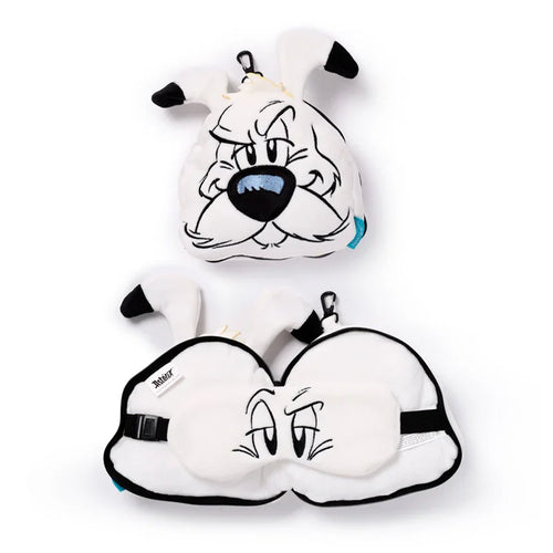 Overhead view of a white plush Idefix (Dogmatix) travel pillow and eye mask set. The top item is the closed round pillow, shaped like a white dog's head with a stern expression, black-tipped ears, and a black carabiner clip. The bottom item is the same travel pillow, unzipped and open to reveal a white eye mask with embroidered sleepy eyes and a black elastic strap.