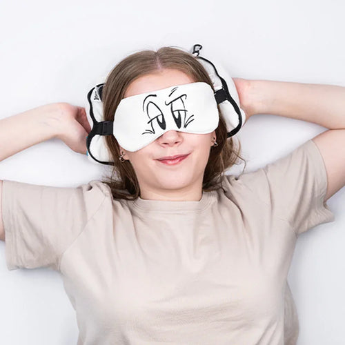 In-context lifestyle shot from above: A smiling woman wearing a white plush travel eye mask with cartoon eyes and a matching round travel pillow.