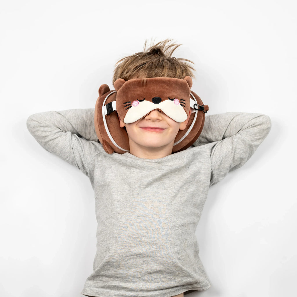 Overhead lifestyle shot of a child wearing a brown otter-themed travel pillow and eye mask. The eye mask, featuring embroidered sleeping eyes and pink cheeks, covers their eyes, while the U-shaped neck pillow supports their head. The child lies comfortably on a white background with hands behind their head.