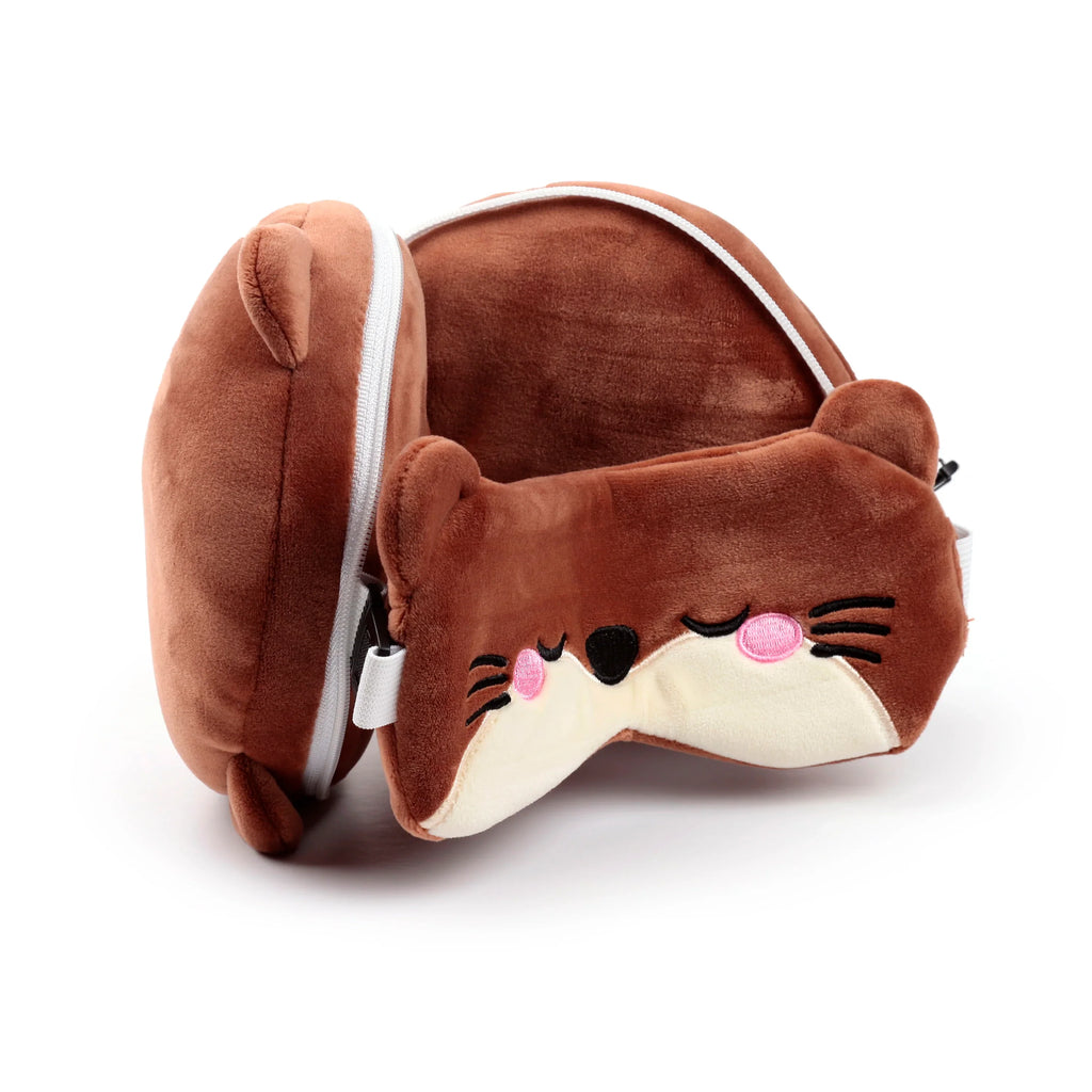 Angled view of a brown plush travel pillow in otter character form, with a white zip, alongside a matching otter-face eye mask featuring embroidered closed eyes, pink cheeks, and black whiskers.
