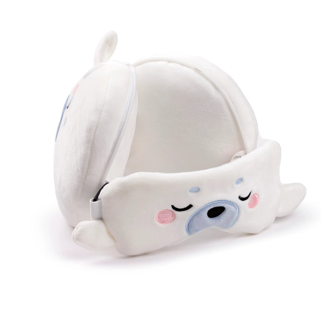Angled view of a white plush Kai the Seal travel pillow and matching eye mask. The eye mask, featuring embroidered sleepy eyes, a blue nose, and pink cheeks, is shown partially stored within the unzipped pillow.