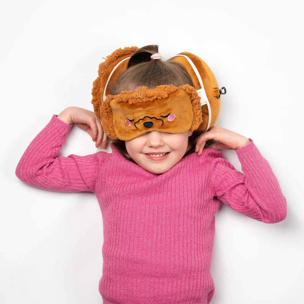 Overhead lifestyle shot of a smiling child in a pink top, wearing a brown toy poodle travel pillow and eye mask set on a white background.