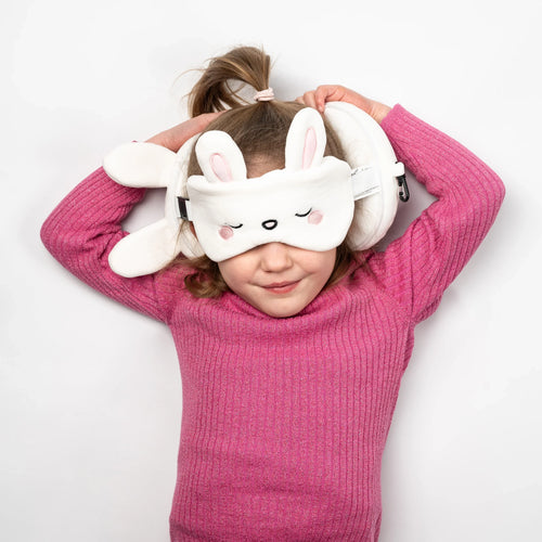 Overhead view of a child wearing a white rabbit travel pillow and eye mask set, featuring embroidered sleeping eyes, pink nose, and cheeks, with stand-up ears.