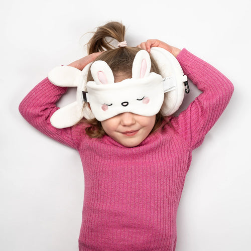 Overhead view of a child wearing a white rabbit travel pillow and eye mask set, featuring embroidered sleeping eyes, pink nose, and cheeks, with stand-up ears.