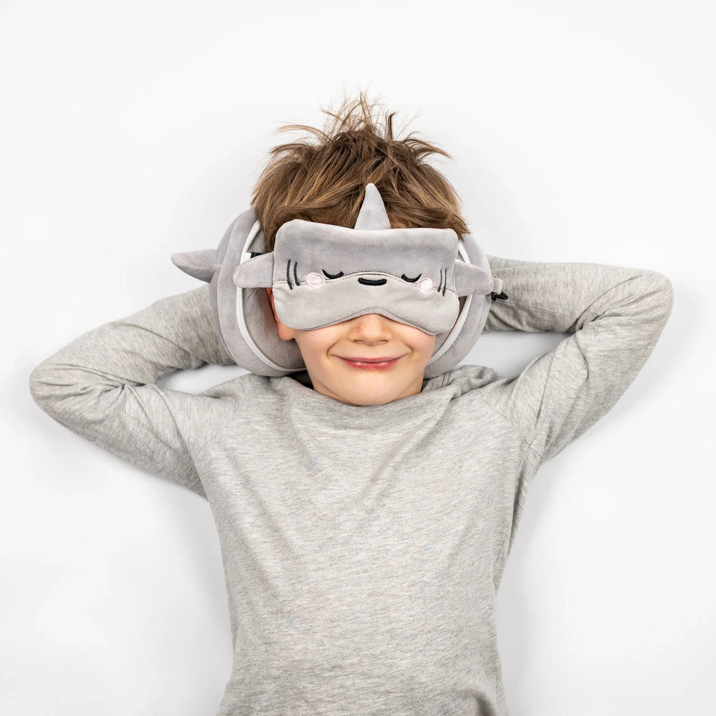 In-context lifestyle shot of a smiling child lying down, wearing a grey Archie the Shark travel pillow and matching eye mask, with the eye mask featuring embroidered closed eyes.