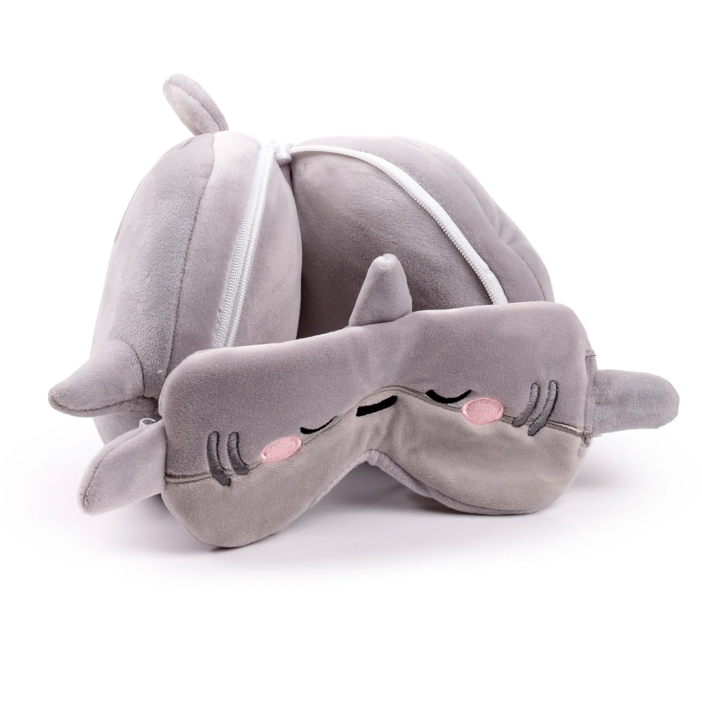 Angled view of a grey plush shark-themed travel pillow and matching eye mask. The eye mask displays embroidered sleeping eyes and pink cheeks, whilst the U-shaped pillow features small fin details and a visible white zipper.