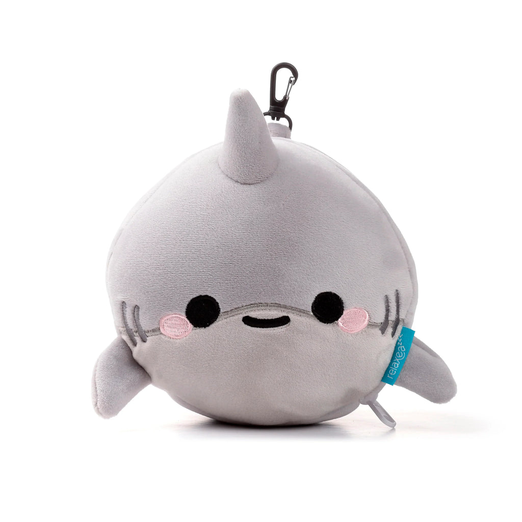 A soft grey plush cartoon shark travel pillow, depicted with embroidered black eyes, a smiling mouth, and pink cheeks. It features small fins and a black carabiner clip at the top.