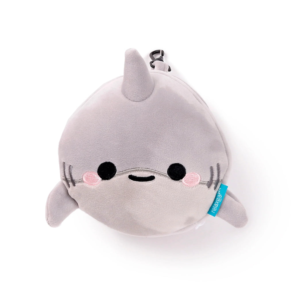 Front view of a grey plush shark travel pillow and eye mask, featuring embroidered black eyes, a small black mouth, pink cheek details, and gill lines. A black carabiner clip is attached at the top, with a small blue fabric tag on its side.