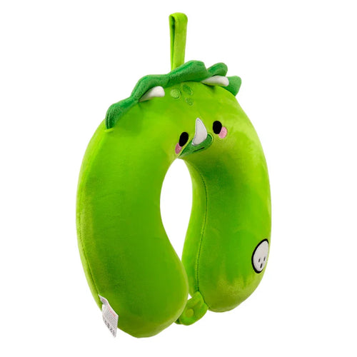 A plush green dinosaur memory foam travel pillow is shown from an angled perspective, hanging by its loop. It features a white horn, black eyes, pink cheeks, green spines along the top, and a small skull emblem on its side.
