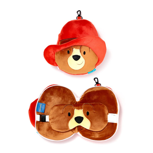 Overhead view of a Relaxeazzz Paddington Bear travel pillow and eye mask set. The top item is the compact plush bear's head with a red hat. The bottom item shows the unfolded U-shaped travel pillow with an integrated eye mask featuring the bear's face and red hat, plus an adjustable strap and clip.