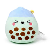 Slightly angled view of a soft, light green and blue bubble tea plush toy, featuring an embroidered happy face with black eyes, a smiling mouth, and pink cheeks. Brown circular 'tapioca pearls' are visible on the green body, with a small yellow straw detail on the blue top.