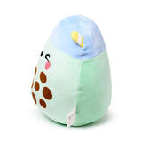 Angled view of a matcha bubble tea plush toy with a light green body, light blue top, and a yellow and white striped straw. It features an embroidered face with closed eyes, a pink tongue, and brown tapioca pearls, with a white label at the bottom.