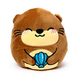 Front view of a brown and cream Squidglys plush toy otter with a smiling embroidered face, holding a blue shell.