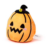 Front-angled view of an orange pumpkin plush toy with black vertical stripes, wearing a black witch's hat with a purple ribbon and yellow buckle. The plush has an embroidered jack-o'-lantern face with triangular eyes, a jagged mouth, and pink rosy cheeks.