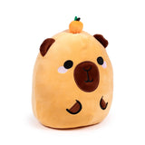 Angled view of a light brown capybara plush toy with embroidered black eyes, dark brown nose and mouth, pink cheek blush, and small brown ears. A tiny orange fruit with green leaves rests on its head.