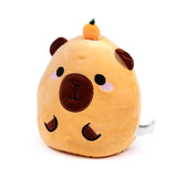 Front view of a Squidglys Duke the Capybara plush toy, light brown, featuring embroidered facial details, pink cheek blush, small brown ears, and a miniature orange fruit on its head.