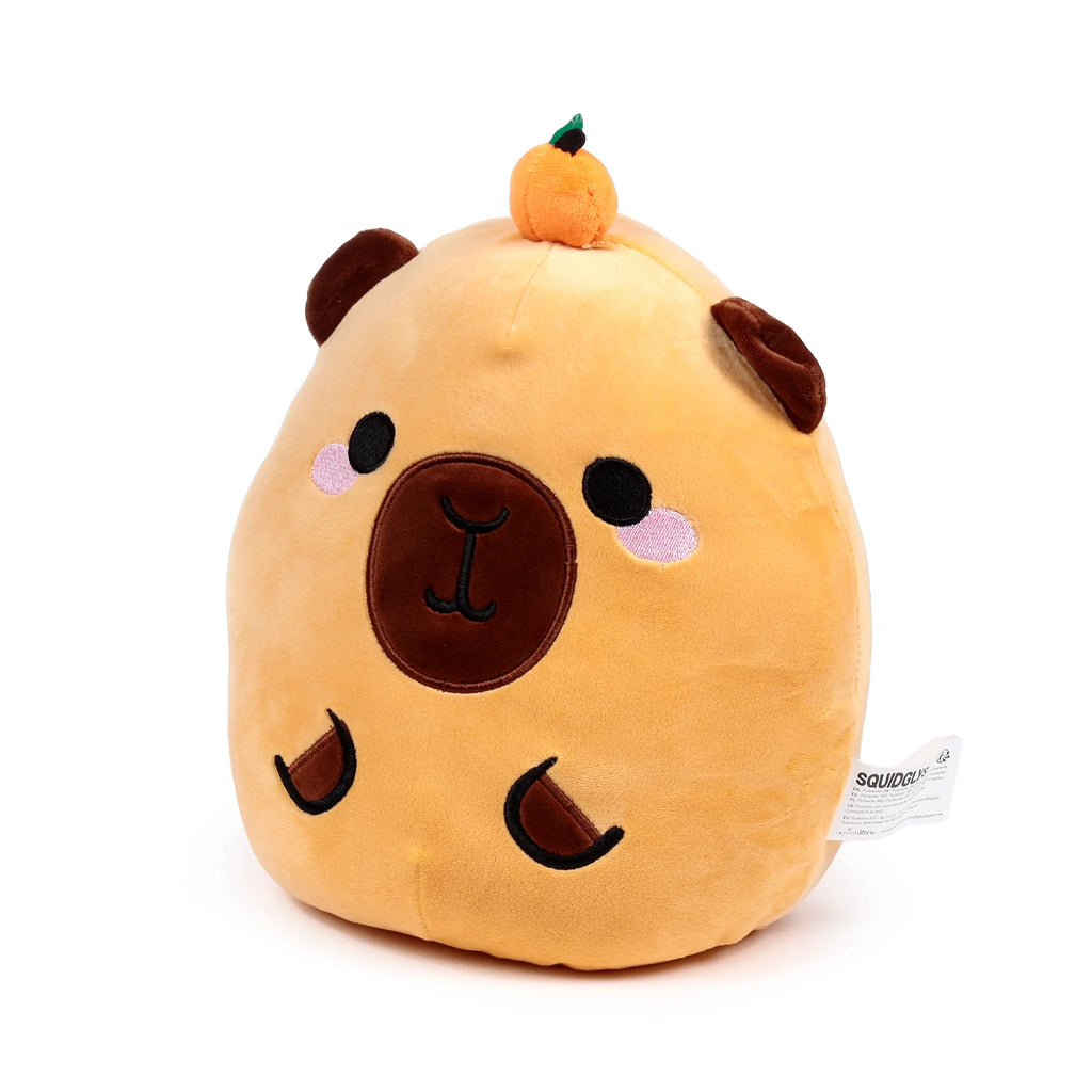 Front view of a Squidglys Duke the Capybara plush toy, light brown, featuring embroidered facial details, pink cheek blush, small brown ears, and a miniature orange fruit on its head.