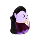 Angled view of a Spooky Dracula plush toy with a lavender face, embroidered closed eyes, a subtle smile, a black top of head, a dark purple body, and a black cape.
