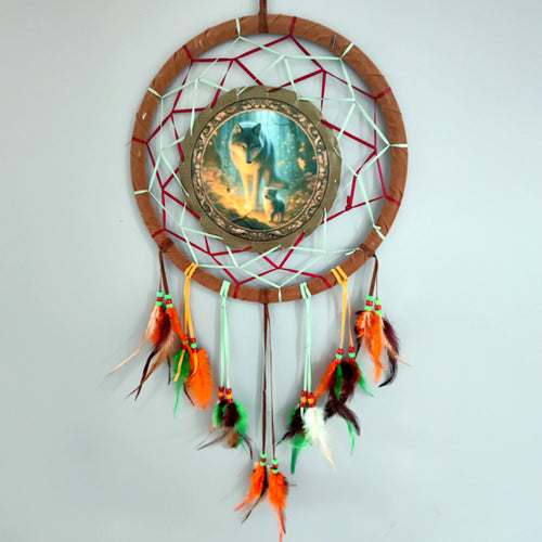 Dreamcatcher with a brown-wrapped hoop, red and mint green woven web, and a central circular illustration of a grey wolf and cub in a glowing forest scene. It features hanging strands adorned with colourful orange, green, brown, and white feathers, accented with red and green beads.