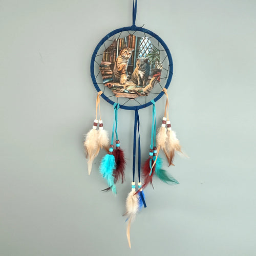 Front view of a navy blue dreamcatcher with a central illustration of two cats, a tabby and a grey, surrounded by books and a magnifying glass, featuring hanging multicoloured feathers and beads.