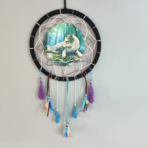 Front view of a black dreamcatcher with a peach-coloured ribbon web, featuring a central circular image of a white wolf and a small fairy in a forest. It has decorative blue, purple, and brown tassels and feathers with beads hanging below.