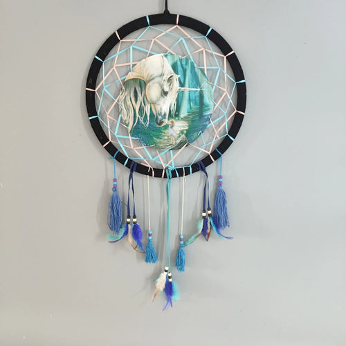 Front view of a dreamcatcher with a black hoop, light blue and pink woven webbing, a central illustration of a white unicorn and a fairy in a forest, and hanging blue tassels with feathers and beads.