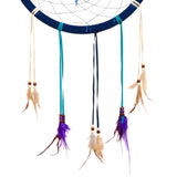 Close-up of the lower section of a blue dreamcatcher, with a fabric-wrapped hoop, blue web, and hanging teal, dark blue, and cream ribbons adorned with wooden beads and natural and purple feathers.