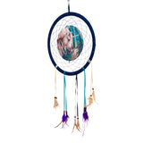 Front view of a dark blue dreamcatcher featuring a round central illustration of a white unicorn and a fairy in a magical forest. It has intricate webbing and hanging teal, dark blue, and cream ribbons adorned with wooden beads, brown, and purple feathers.