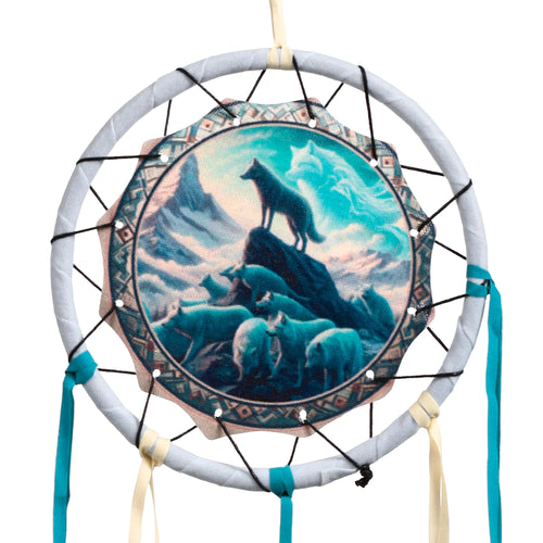 Front view of a dreamcatcher featuring a central illustration of a black wolf on a rocky peak, overlooking a pack of wolves in a blue and teal mountainous landscape. The dreamcatcher has a light grey fabric-wrapped hoop, black webbing, and hanging turquoise and cream ribbons.