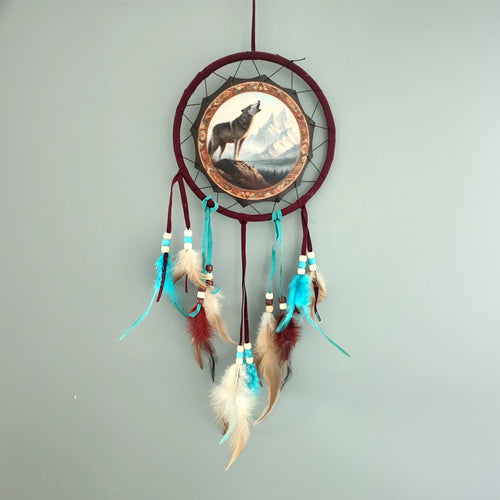 Front view of a small dreamcatcher with a dark red hoop. The central disc depicts a howling mountain wolf against a backdrop of rocky peaks and blue sky. Strands adorned with cream and light blue beads, and a mix of brown, cream, and turquoise feathers hang below.