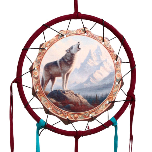 Front view of a small dreamcatcher, depicting a howling grey wolf on a rock with snowy mountains and a forest. It features a circular, deep red velvet-wrapped frame and hanging red and turquoise fabric strips.