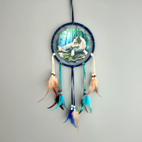 Blue dreamcatcher with a central image of a greyish wolf and a small fairy in a forest setting. It features hanging fawn, turquoise, and dark blue feathers, complemented by wooden beads and colourful ribbons.