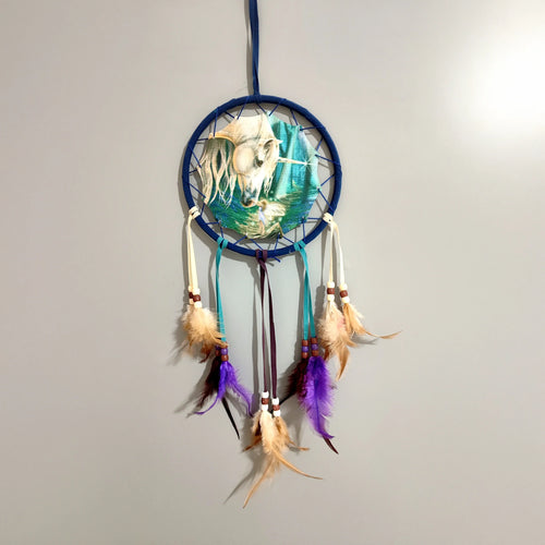 Front view of a small blue dreamcatcher, featuring a central illustration of a unicorn and a fairy, with dangling purple and tan feathers and wooden beads.