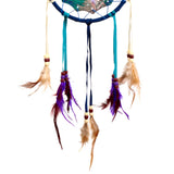 Close-up of a dreamcatcher's lower section, featuring a dark blue fabric-wrapped hoop with a colourful printed pattern. Teal and dark blue fabric strands hang down, adorned with cream and reddish-brown beads, from which dangle a variety of purple, maroon, and light brown feathers, against a white background.