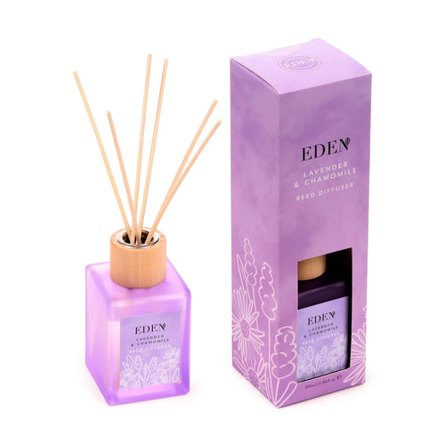 Angled view of a translucent lavender-purple square reed diffuser bottle, featuring natural reeds and a wooden collar, displayed next to its matching lavender-purple gift box. Both the bottle and the box are adorned with 'Lavender & Chamomile Reed Diffuser' text and delicate white floral illustrations.