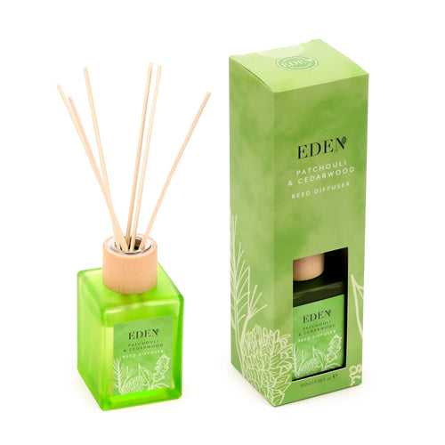 Angled view of a green glass reed diffuser bottle with natural wooden sticks, displayed next to its matching green packaging box. Both the bottle label and box feature 'Eden Patchouli & Cedarwood Reed Diffuser' text and delicate botanical line art.