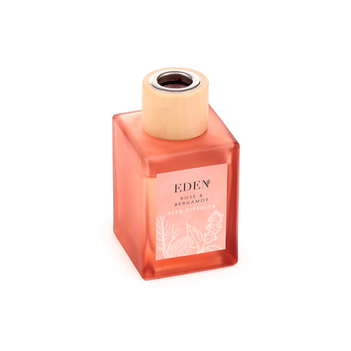 Angled view of a coral square glass Eden Rose & Bergamot reed diffuser bottle, featuring a light pink label with botanical illustrations and a natural wooden stopper.