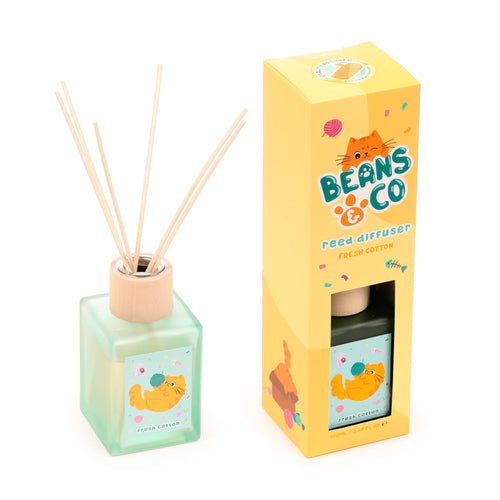 Angled view of a square, translucent light green reed diffuser bottle with a wooden top and several natural reeds, positioned next to its yellow packaging box. Both the bottle and box feature a playful cartoon cat design, 'Beans & Co' text, and 'Fresh Cotton' scent label.