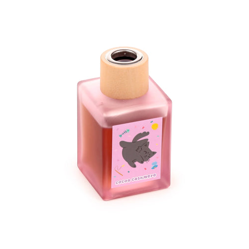 Angled view of a square, frosted pink glass reed diffuser bottle with a light wooden top. A pink label depicting a grey cartoon cat and 'cocoa cashmere' text is visible on the side.