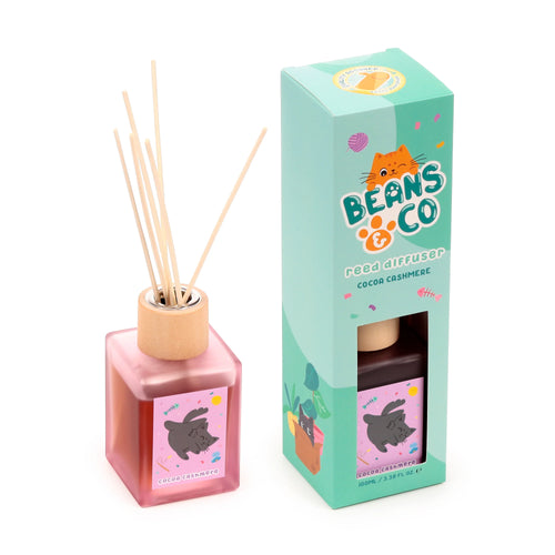 Angled view of a square, translucent pinkish-red reed diffuser bottle with a wooden collar and natural wooden reeds, featuring a label with a grey cat and 'cocoa cashmere' text. Beside it is a mint-green packaging box with an orange cat logo, 'Beans & Co reed diffuser Cocoa Cashmere' text, and a cut-out revealing the product inside.