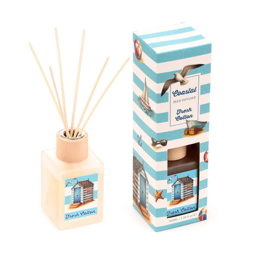 Angled view of a Coastal Fresh Cotton reed diffuser. The square, translucent bottle with a wooden top and reeds has a label showing a beach hut and seagull. Beside it stands the blue and white striped packaging box, adorned with coastal illustrations like a seagull, sailboat, and beach huts, displaying 'Coastal Reed Diffuser Fresh Cotton' text.