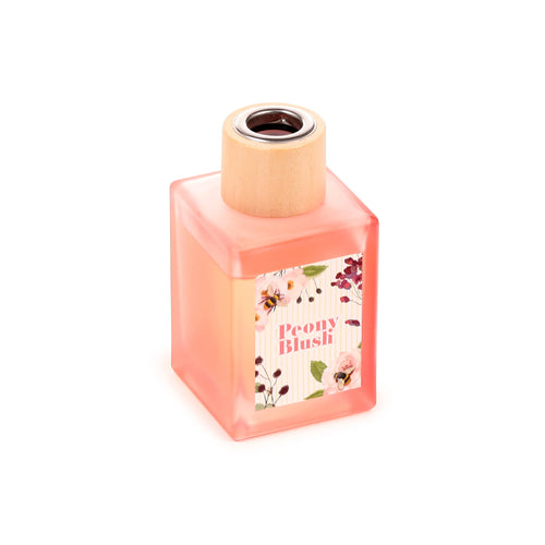 Angled view of a square frosted glass reed diffuser, with peach-pink liquid and a natural wooden collar with a silver-coloured ring. The label features peonies, berries, bees, and 'Peony Blush' text.