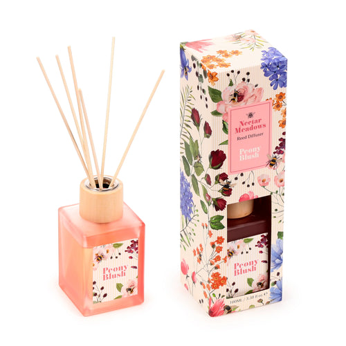 Angled view of a Nectar Meadows Peony reed diffuser, featuring a translucent peach bottle with natural reeds and a 'Peony Blush' floral label. It is placed beside its tall, decorative box, which is adorned with matching colourful floral and bee designs, and displays 'Nectar Meadows Reed Diffuser Peony Blush' text.