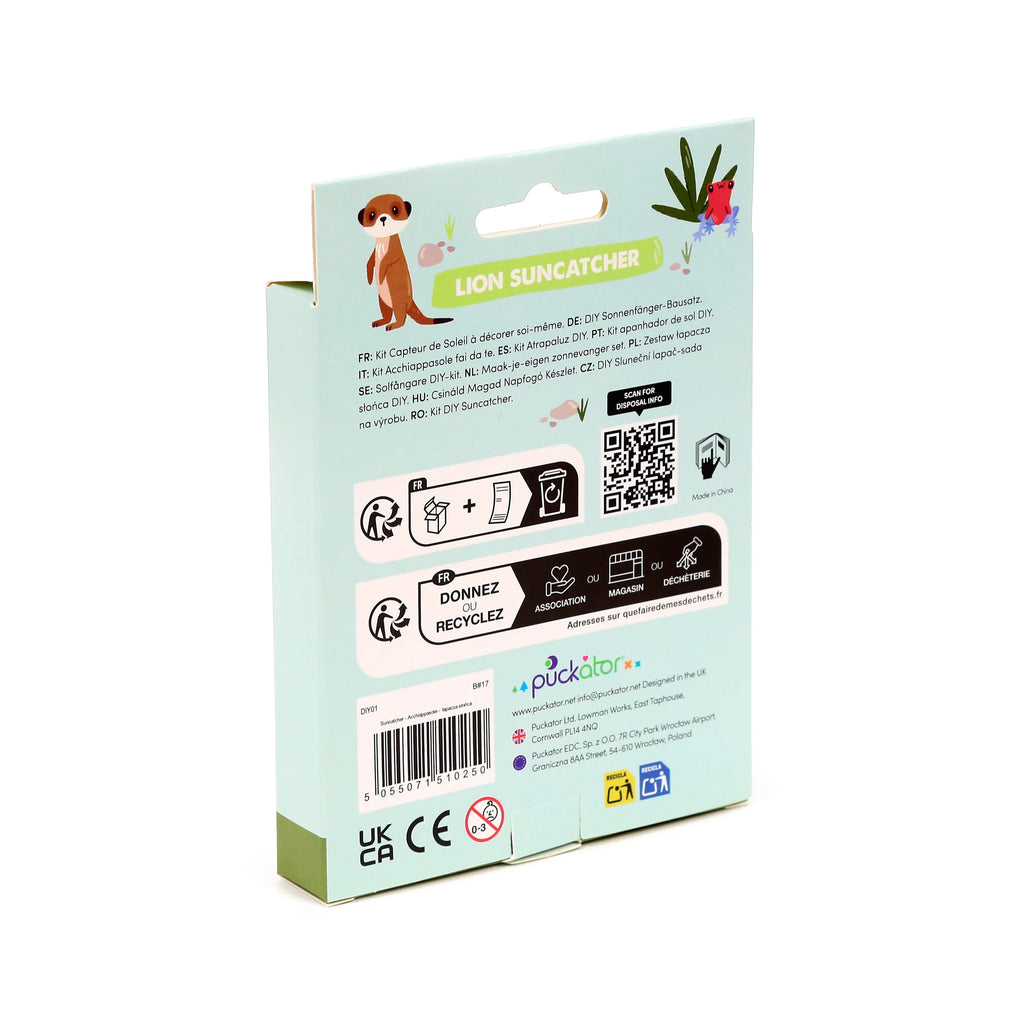 Angled view of the back of a DIY Suncatcher kit box for a 'Lion Suncatcher'. The light blue-green packaging features cartoon illustrations of a meerkat and a red frog, multi-language product details, a barcode, safety warnings, and recycling information.