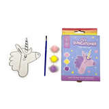 DIY unicorn suncatcher kit components displayed on a white background, featuring an unpainted unicorn outline with a hanging loop, a blue paintbrush, three small pots of pink, yellow, and purple paint, and the kit's purple packaging box.