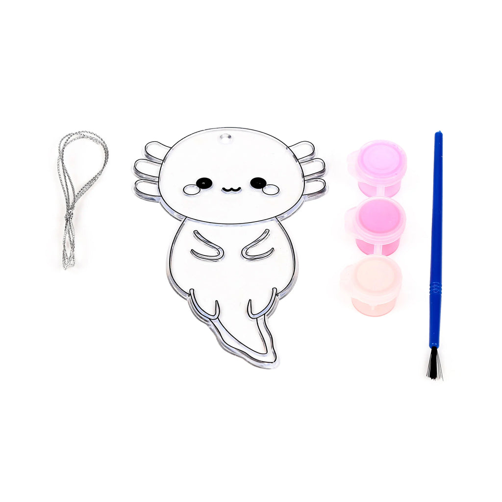 Overhead view of a DIY axolotl suncatcher kit, showing a clear axolotl shape, two pink and one peach paint pot, a blue paintbrush, and silver hanging string.